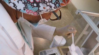Healthcare worker in scrubs, mask, and gloves uses a pipette to dispense liquid into a container in a laboratory.