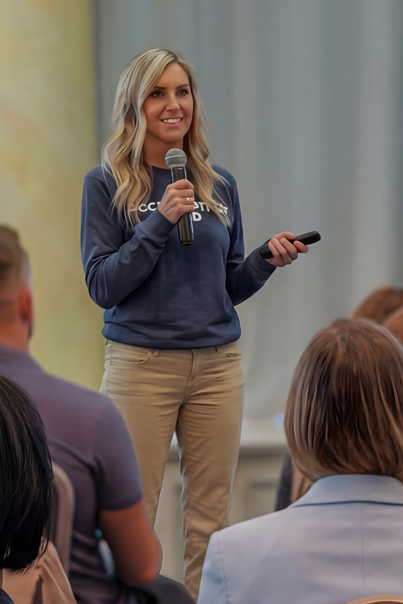 Woman presenter stands at front, holding a microphone and remote, addressing an audience inside a room.