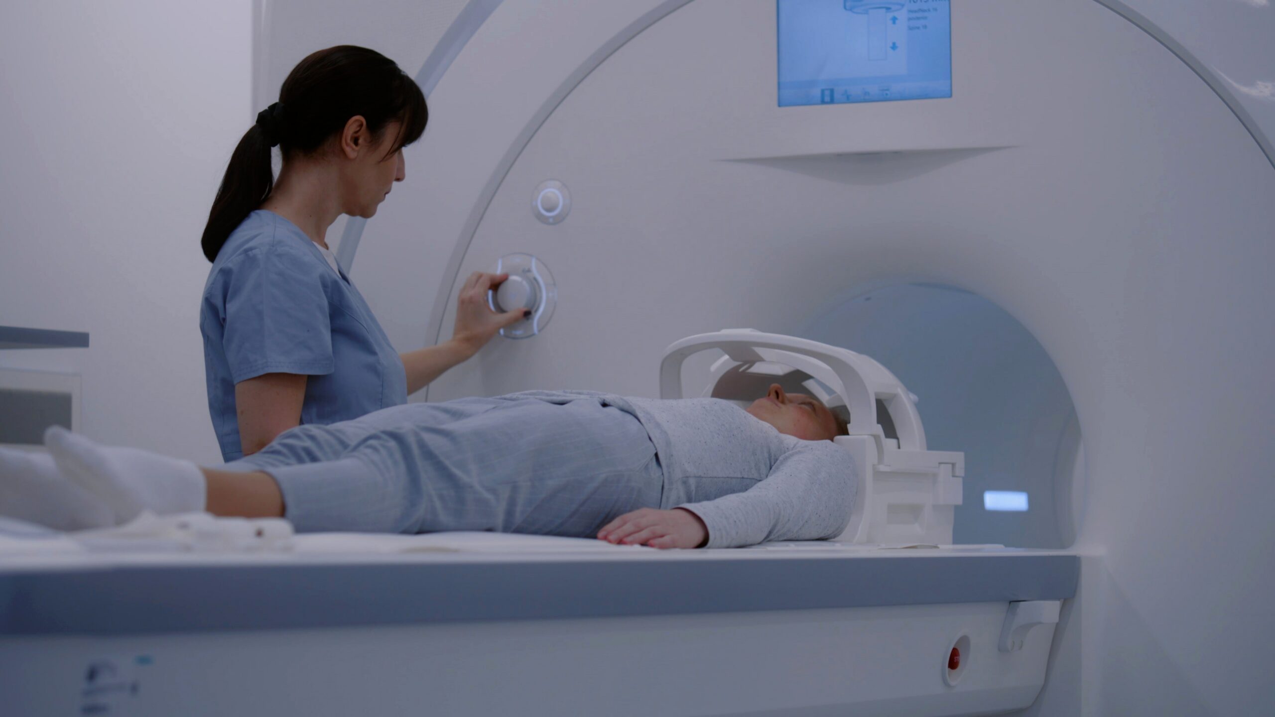 Healthcare worker in blue scrubs adjusts an MRI machine as a patient lies on the scanner bed awaiting imaging.
