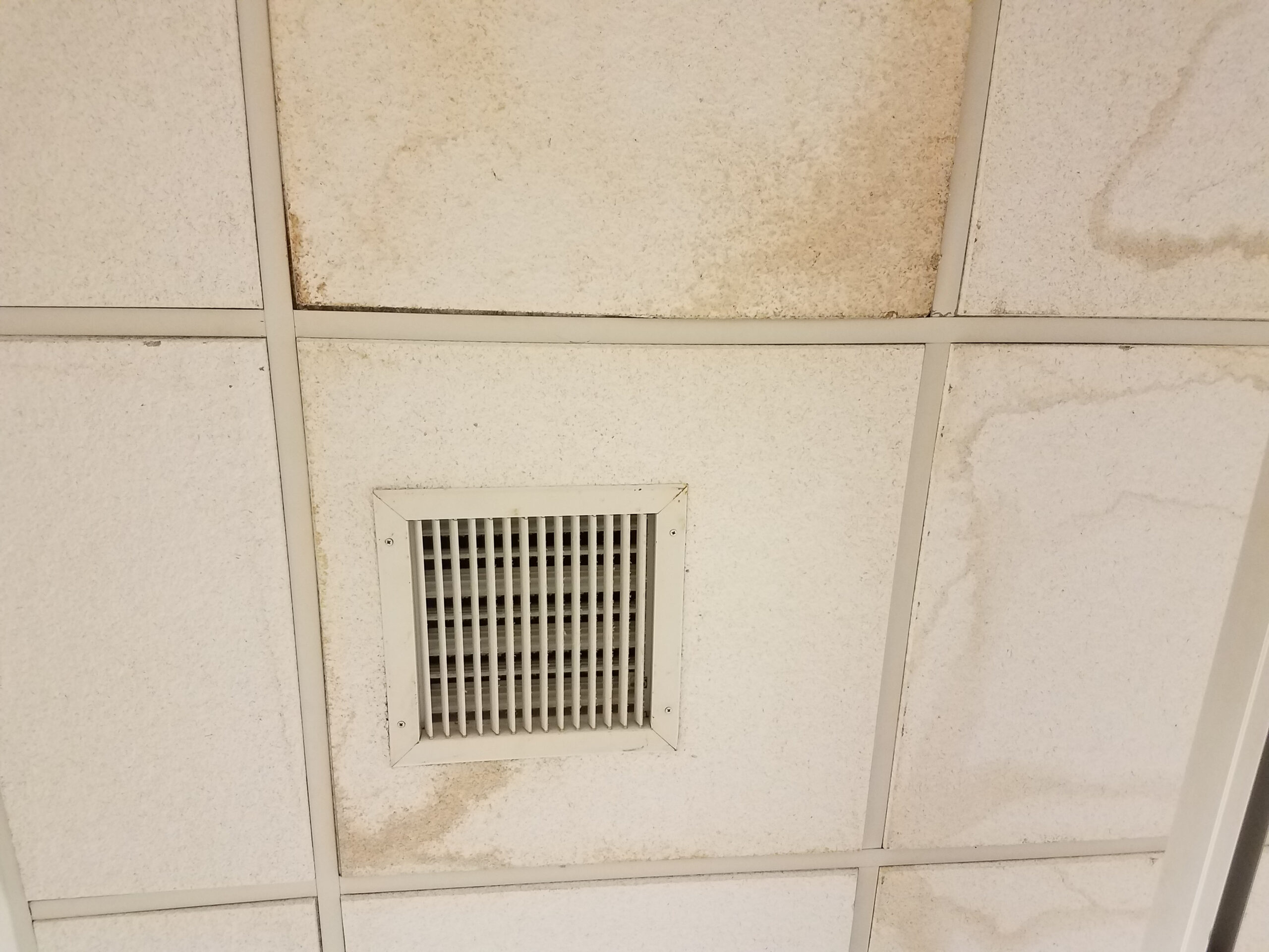 Ceiling vent surrounded by white tiles with brown stains and grime gathering around the edges at the center of the image.