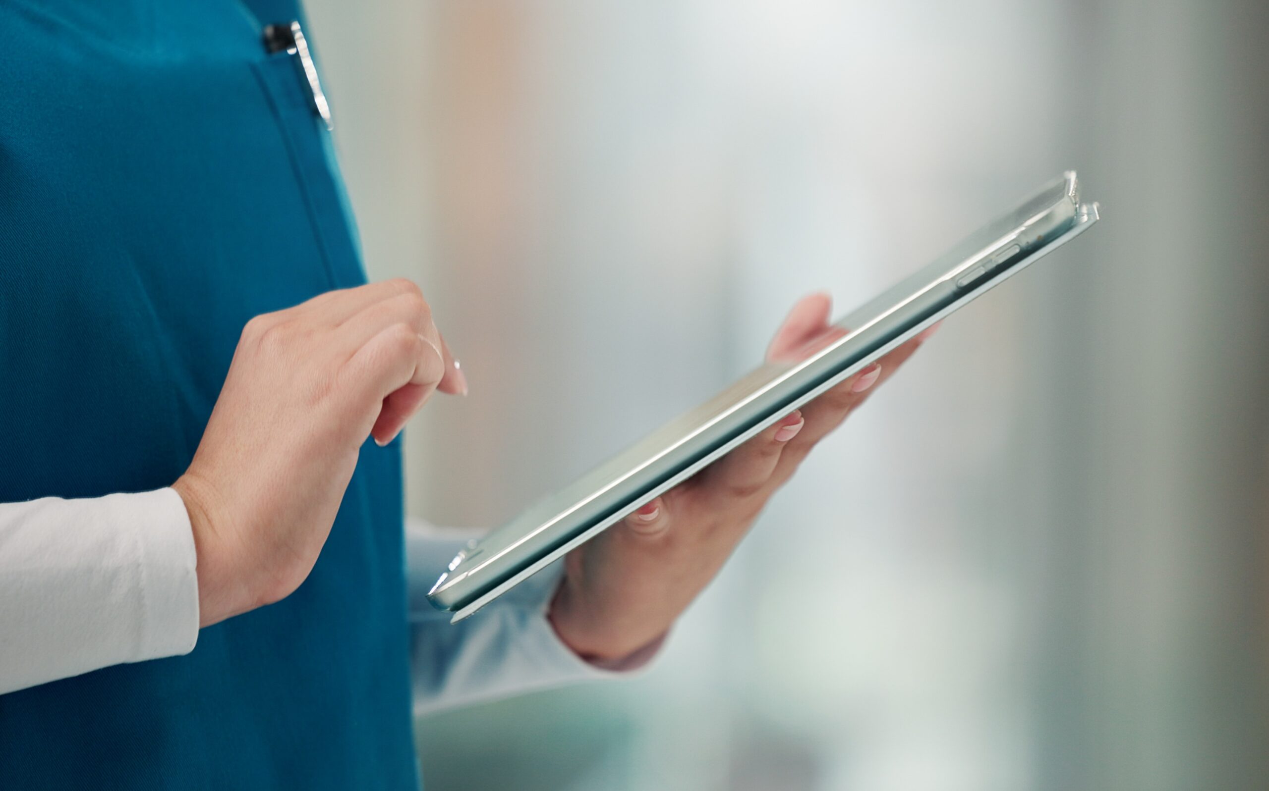 Healthcare professional in teal scrubs using a tablet device in a clinical setting.
