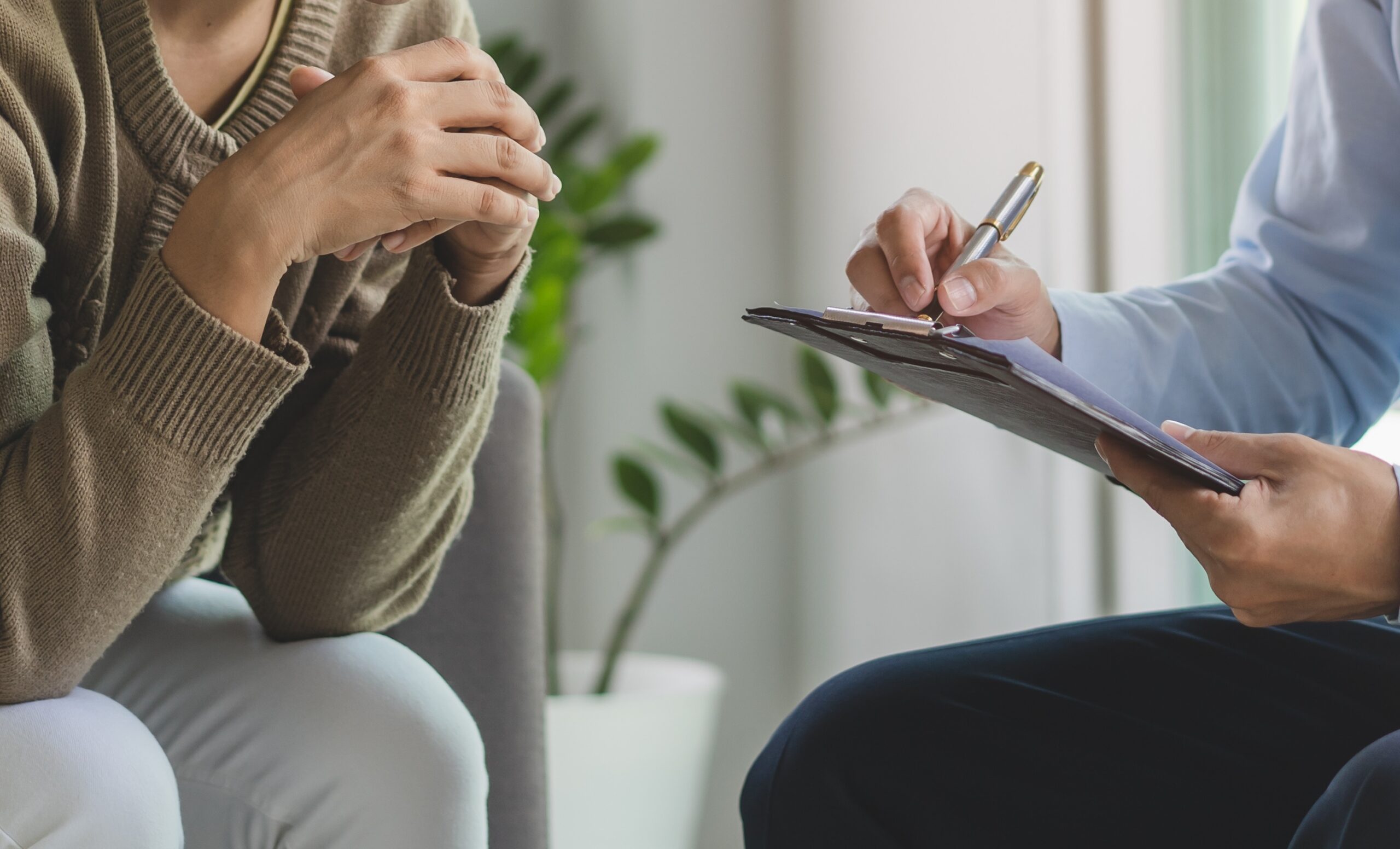 Client with folded hands in a brown sweater while a clinician writes on a clipboard nearby.