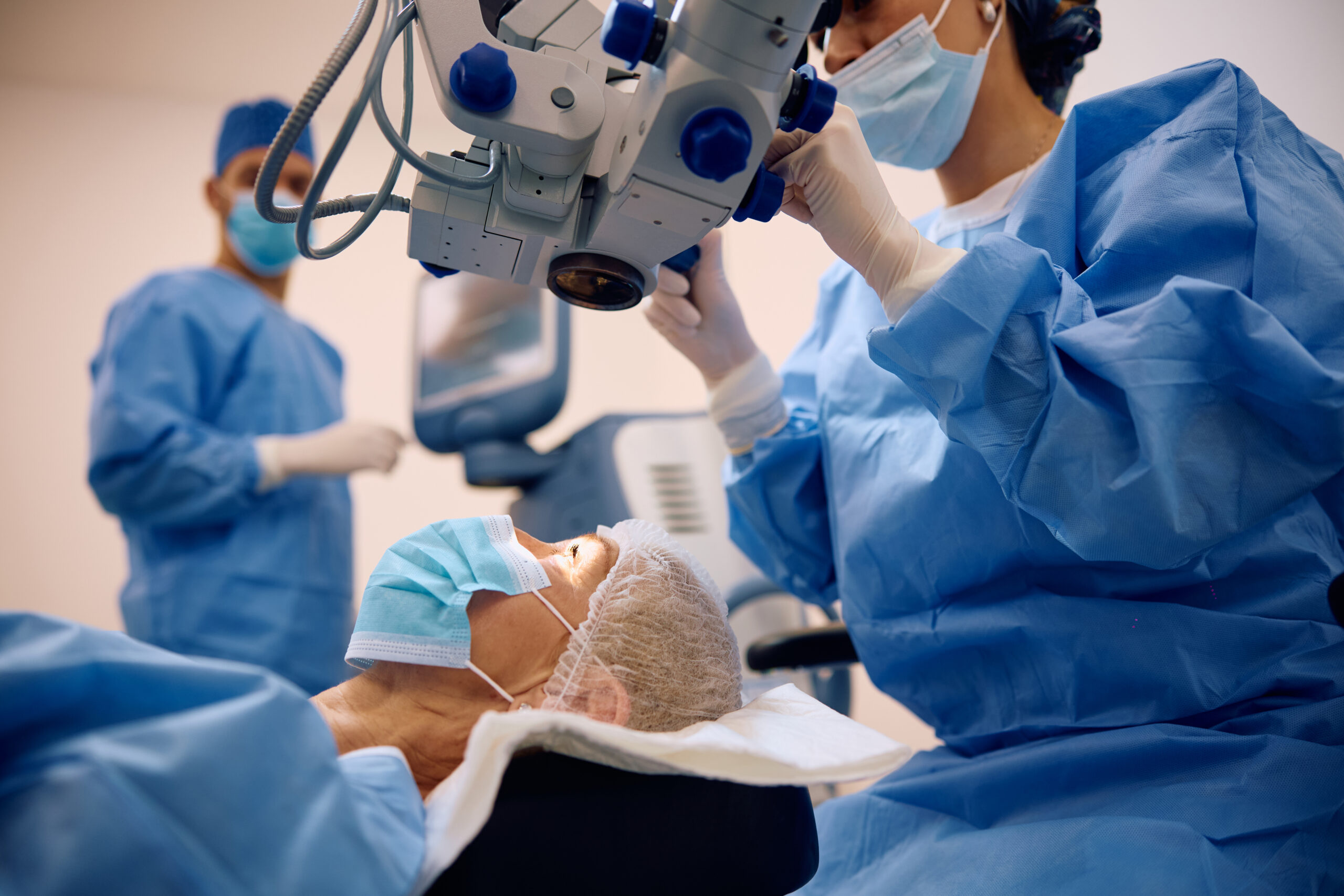 Surgeons in blue scrubs and masks operate around a patient under a medical microscope.