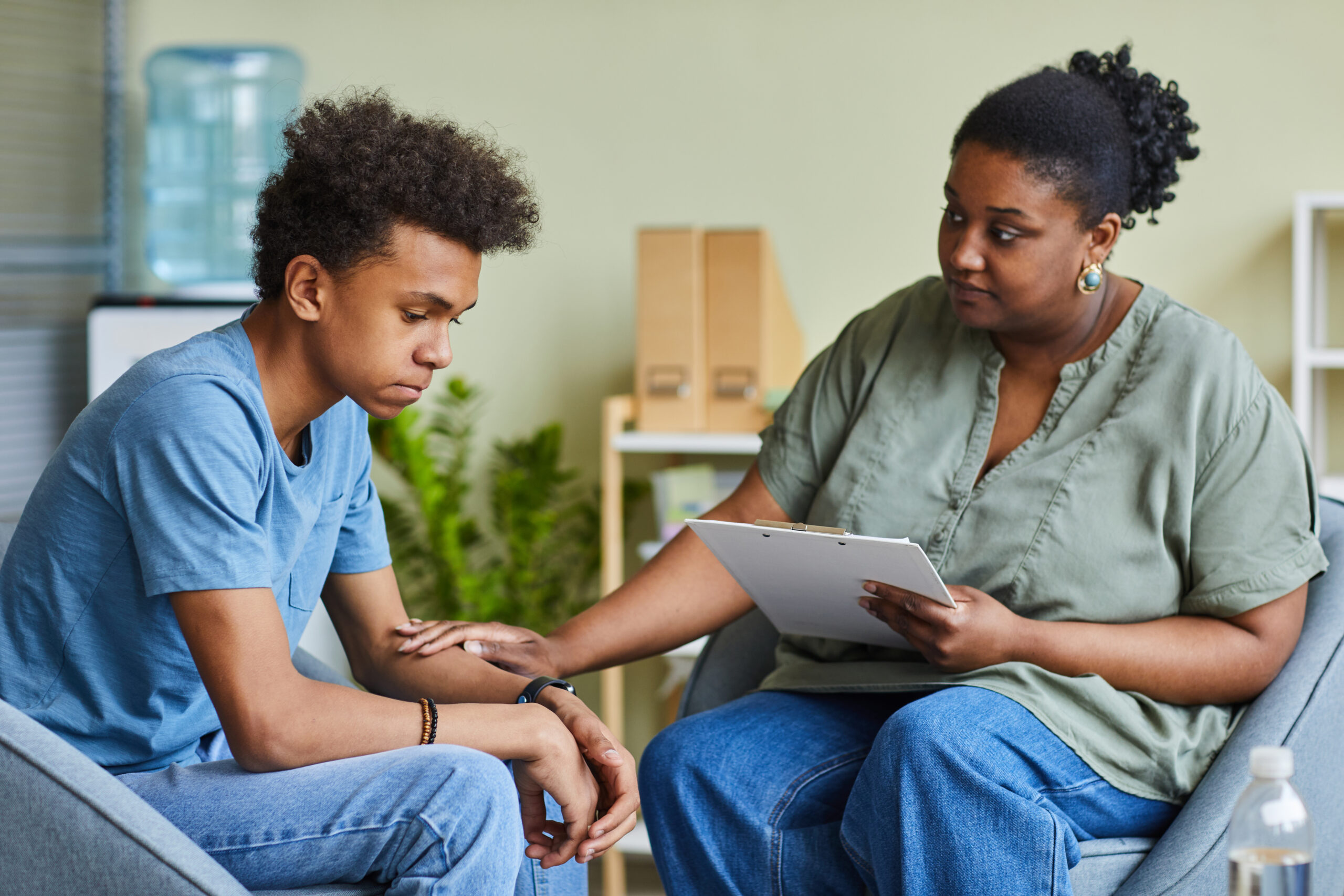 Two adults share a conversation in a clinic-like setting: a woman with a clipboard speaks to and supports a young man who rests his arm on his lap.