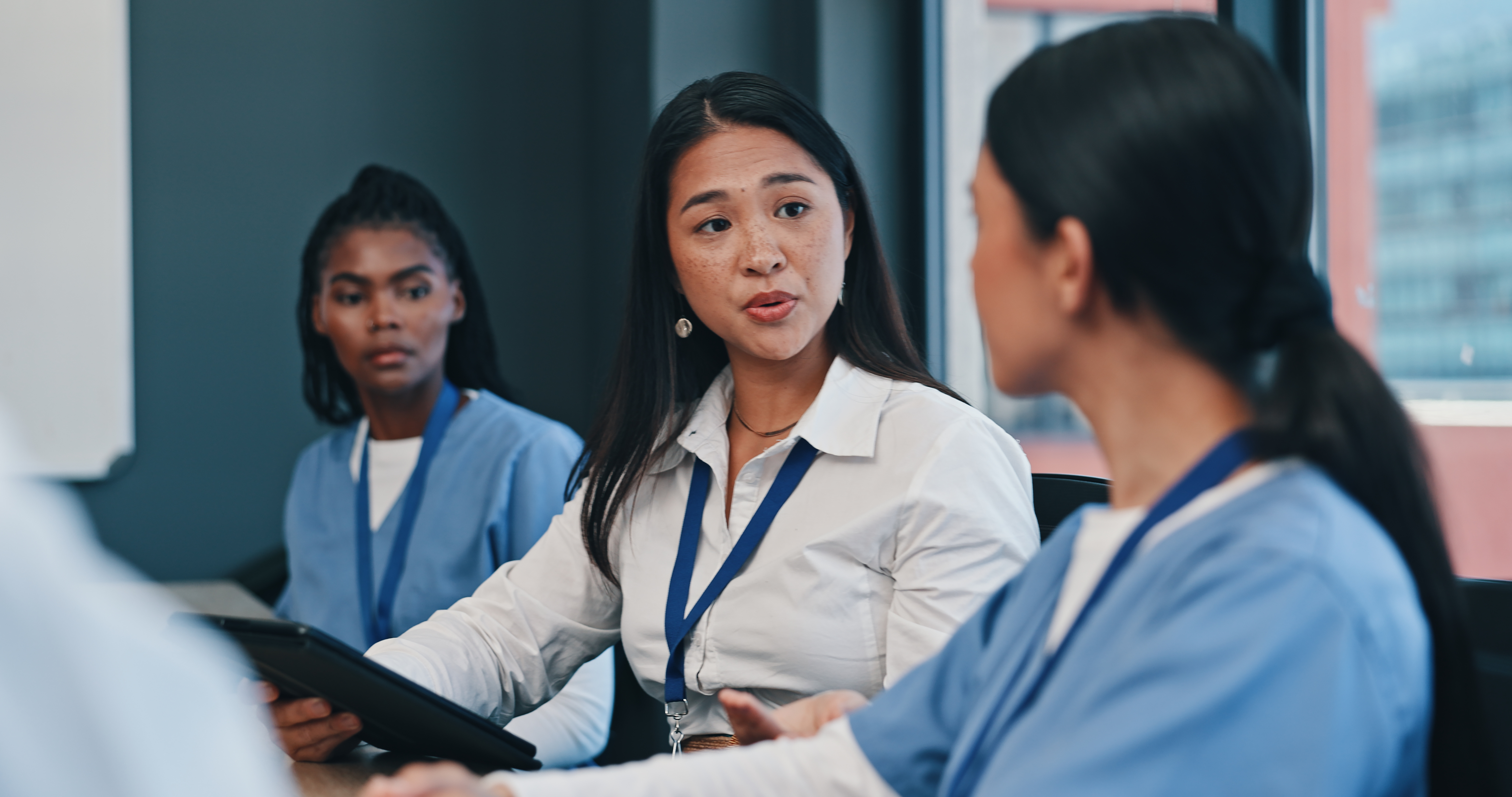 Woman in a white blouse speaks to two colleagues in blue scrubs during a meeting around a table in an office.