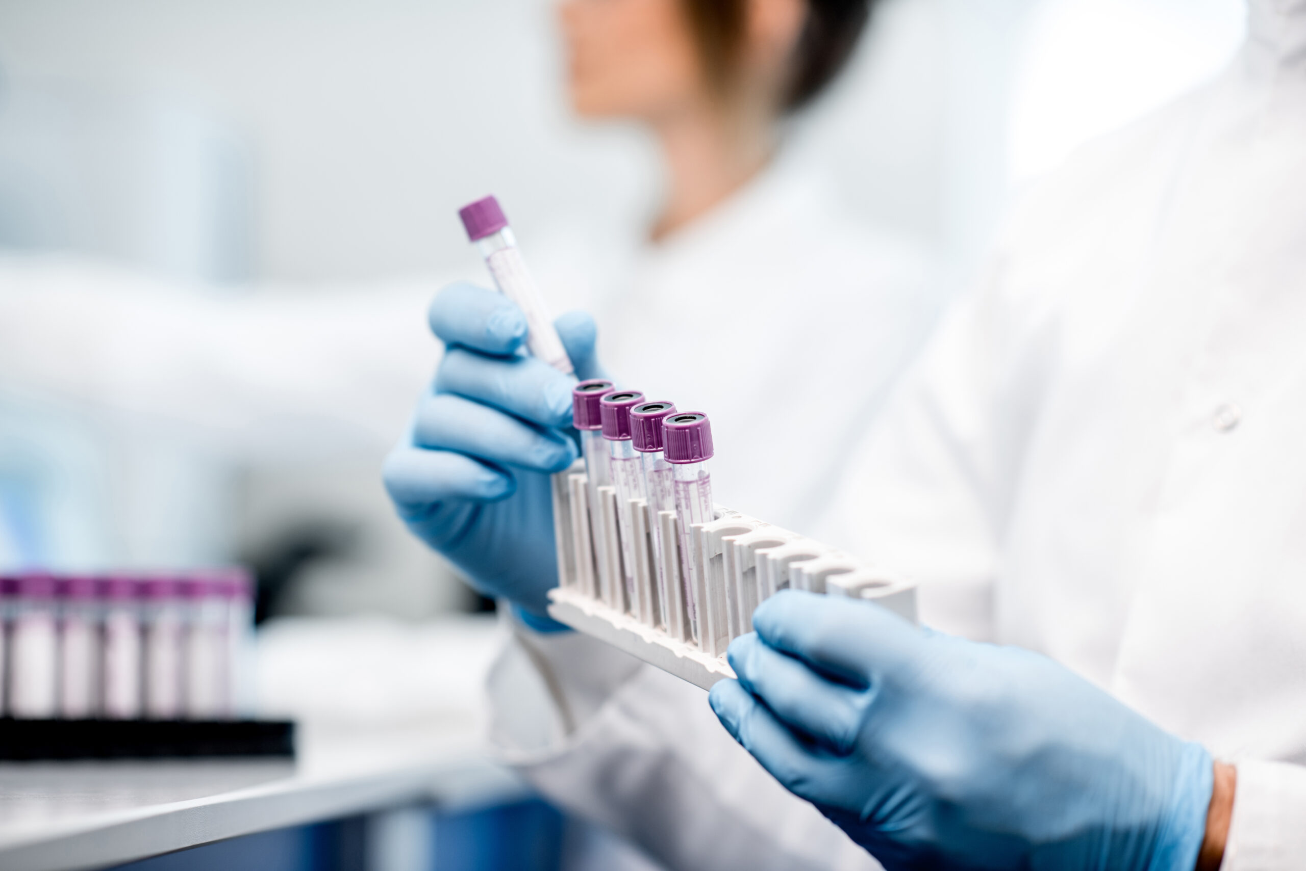 Laboratory technician wearing blue gloves holds a rack of test tubes with purple caps in a lab.