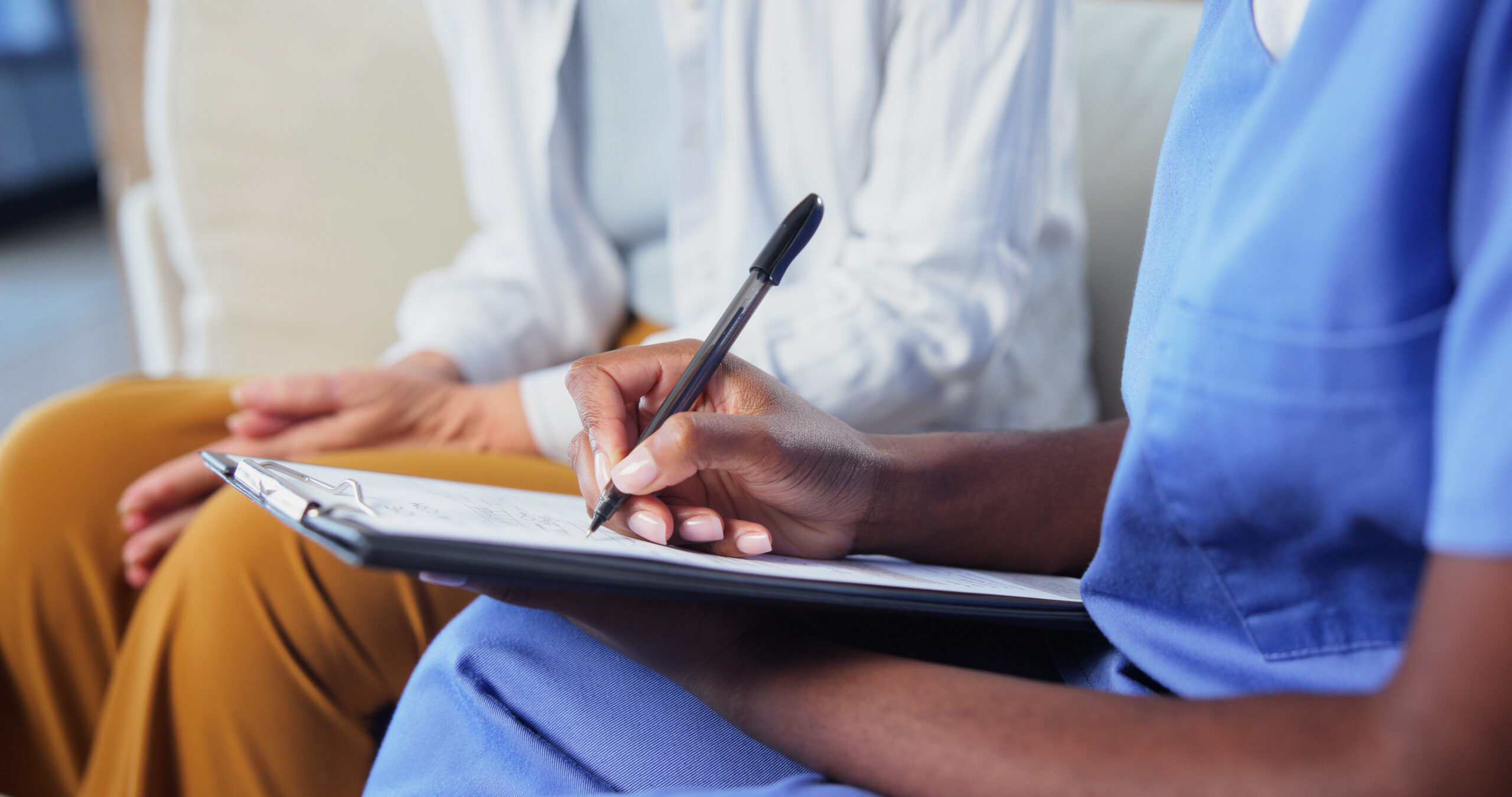 Two people in a clinic setting; one writes on a clipboard while the other holds it nearby.