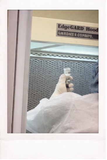 Picture A | ACHC.ORG Gloved hand in protective suit holding a small vial inside a laboratory biosafety hood; EdgeGARD Hood sign visible above.