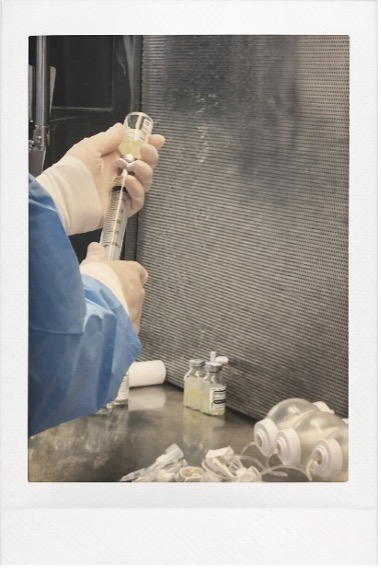 Picture B | ACHC.ORG A lab technician wearing a blue protective gown draws liquid into a syringe at a workbench with vials and syringes nearby.