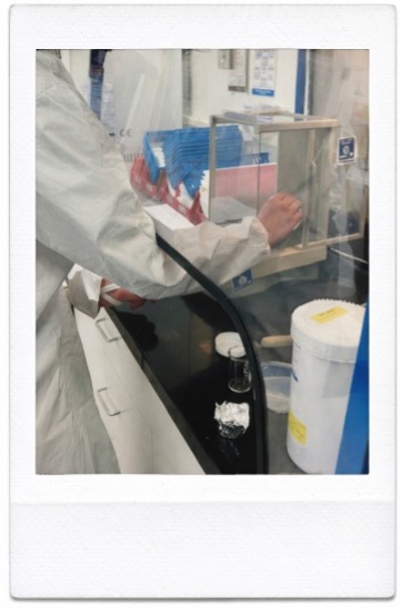 Picture C | ACHC.ORG Lab technician in a white lab coat uses a biosafety cabinet, with blue racks and lab supplies nearby.